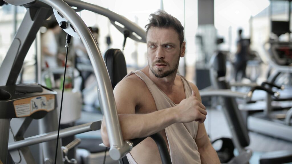 Muscular man resting on gym equipment in a modern fitness center, looking determined.