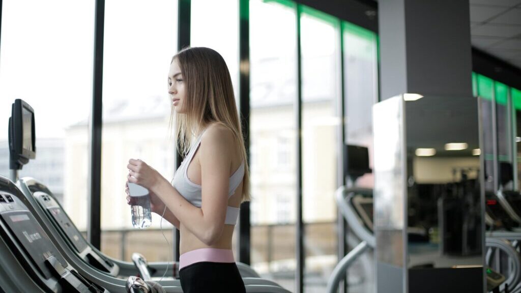Side view of slim young female athlete in activewear standing on treadmill with bottle of fresh water while resting during training in modern light gym